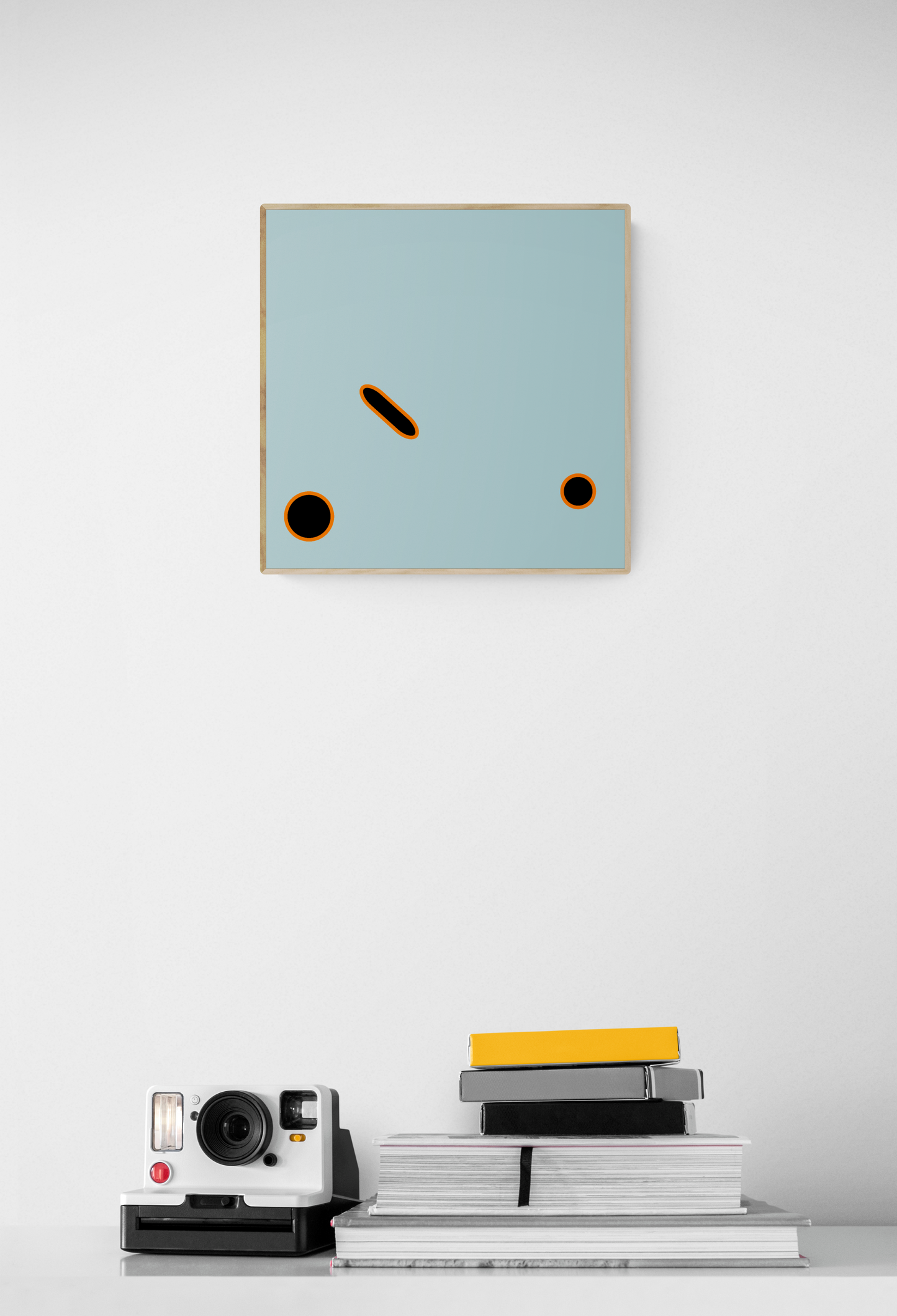 Abstract wall art print with geometric shapes on a white wall, below which are books and a camera.