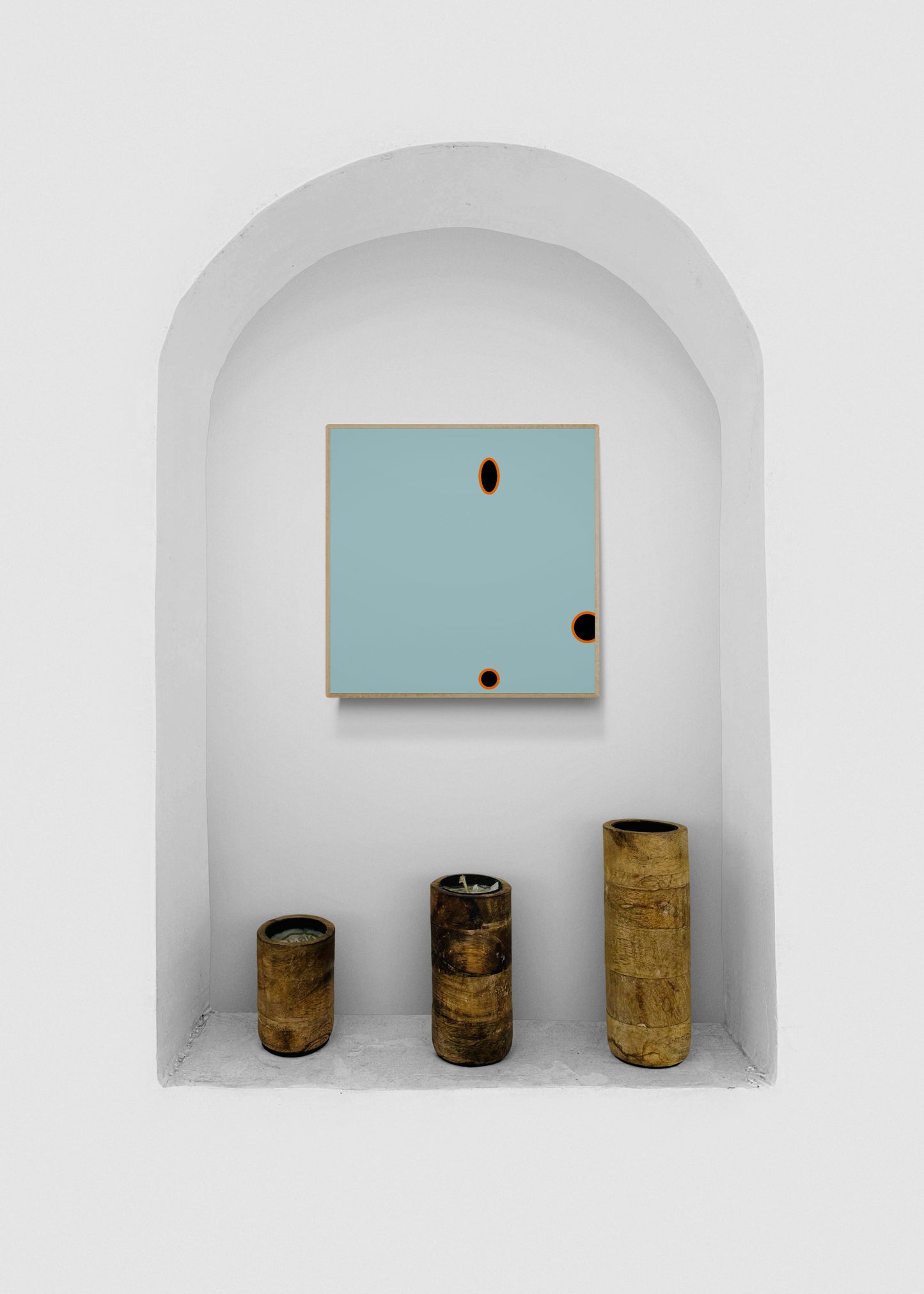 Decorative wall art print with a light blue square and black circular cutouts, displayed in a white alcove with three cylindrical wood objects.