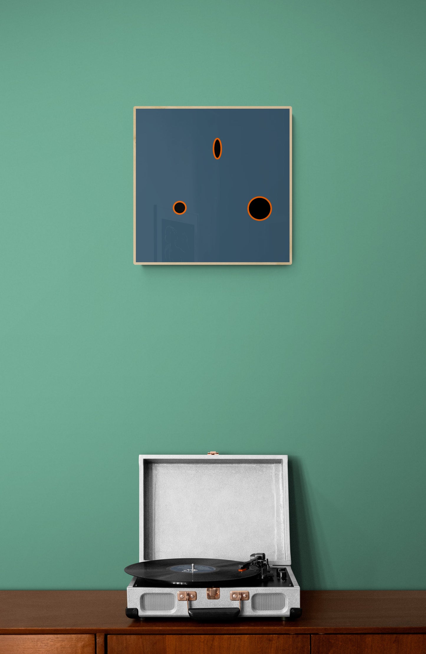 Record player on a wooden surface with a framed grey abstract art print on a green wall.