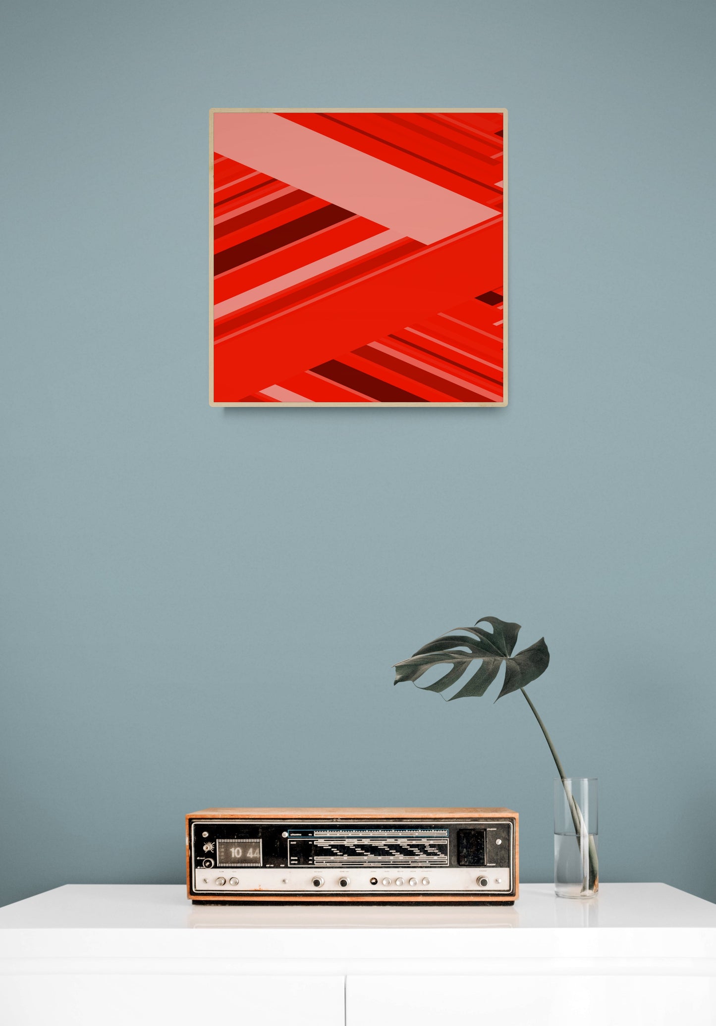 Abstract red and black geometric art print on a gray wall above a white surface with a vintage radio and plant.