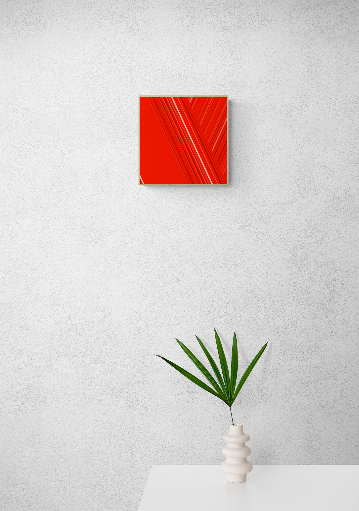 Art print of abstract red design with geometric shapes on a red background on a white wall with a white vase containing green leaves below.
