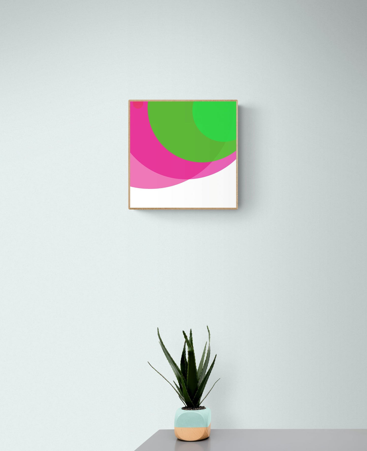 Art print of two overlapping circles, one green and one pink, on a white backgroundwith pink and green shapes on a white wall, accompanied by a potted plant.