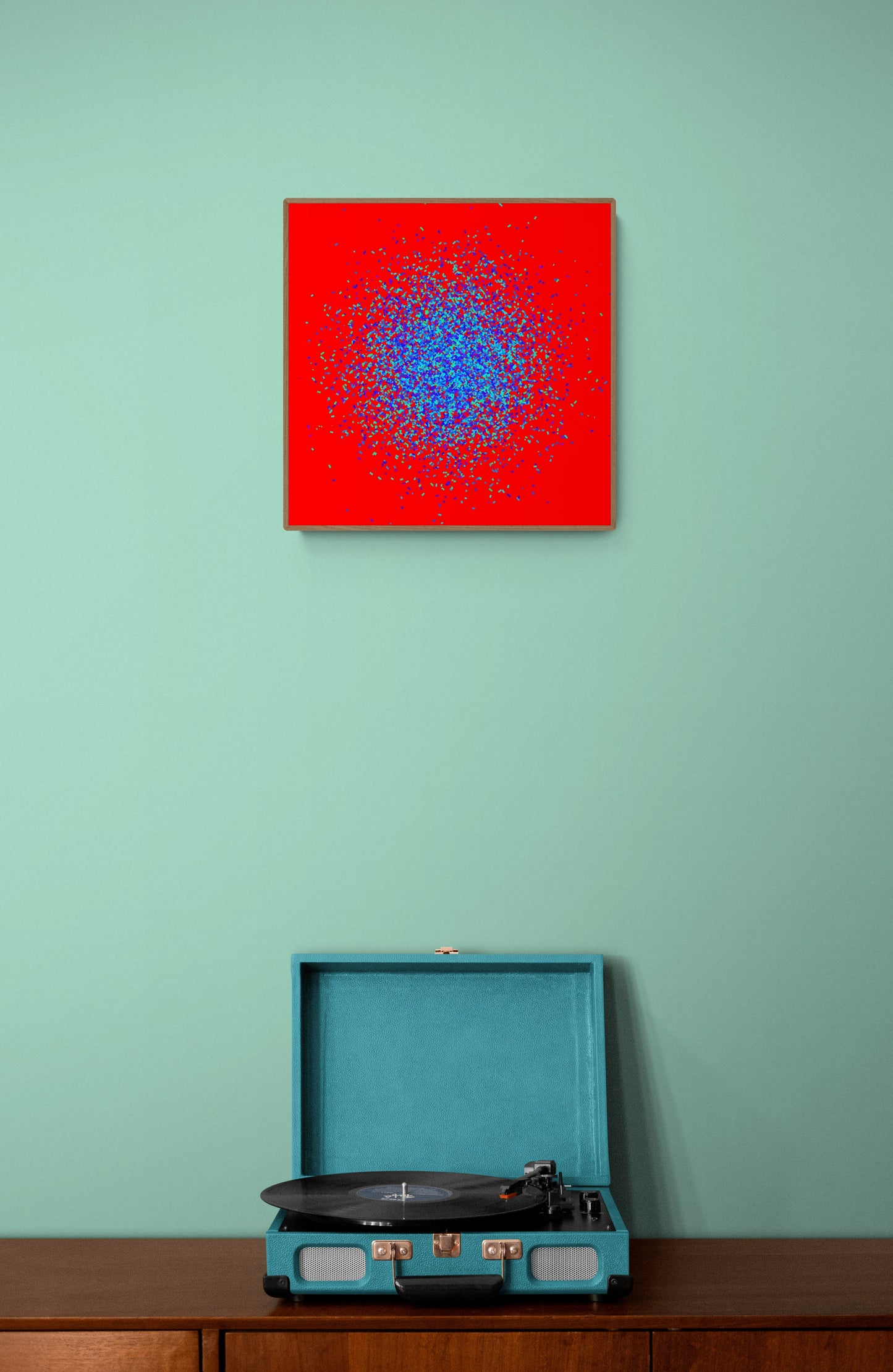 Teal record player on a wooden surface with an abstract patterned art print of blue and green fragments on a red background on the wall.
