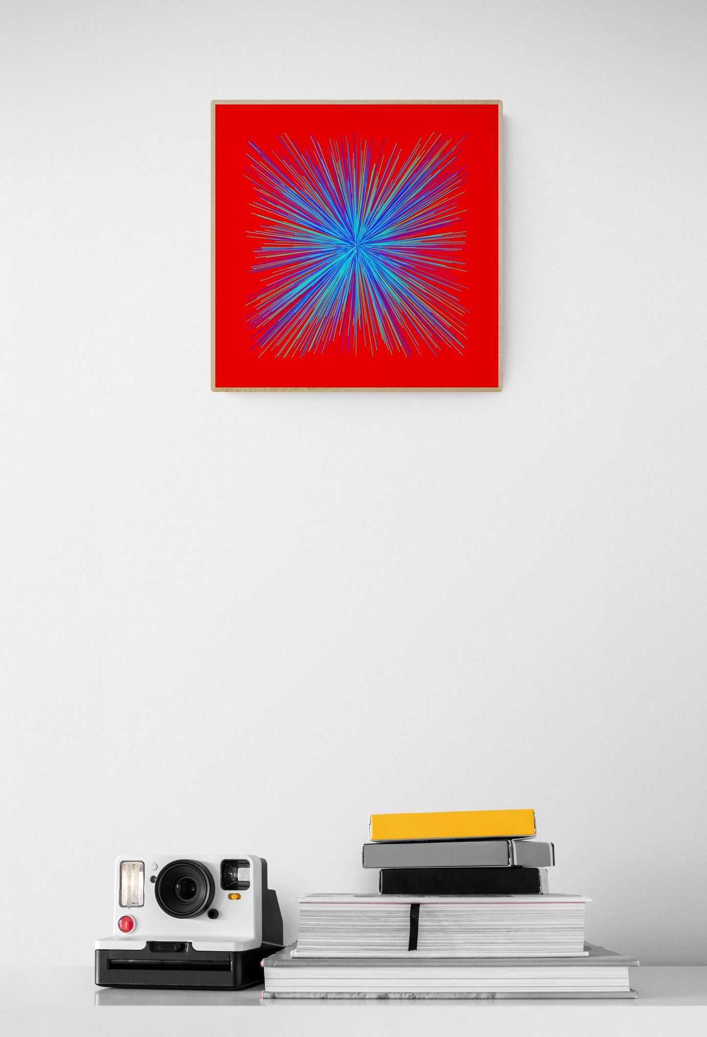 Abstract art print of colorful blue, green radialburst pattern on a red background on a white wall, below which are books and a camera.
