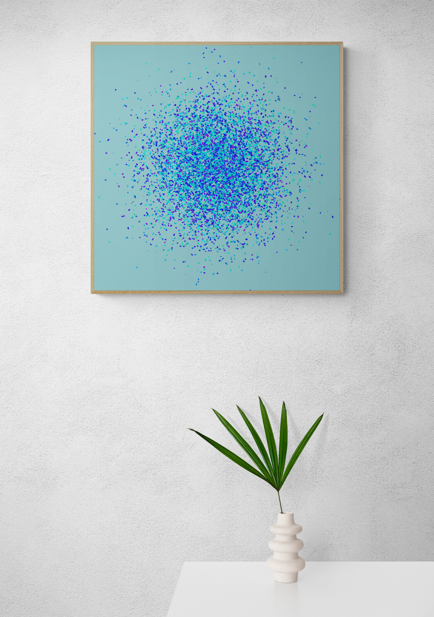 Framed abstract art print of blue, green and purple confetti like circular pattern on a light blue background, placed above a white table with a plant.
