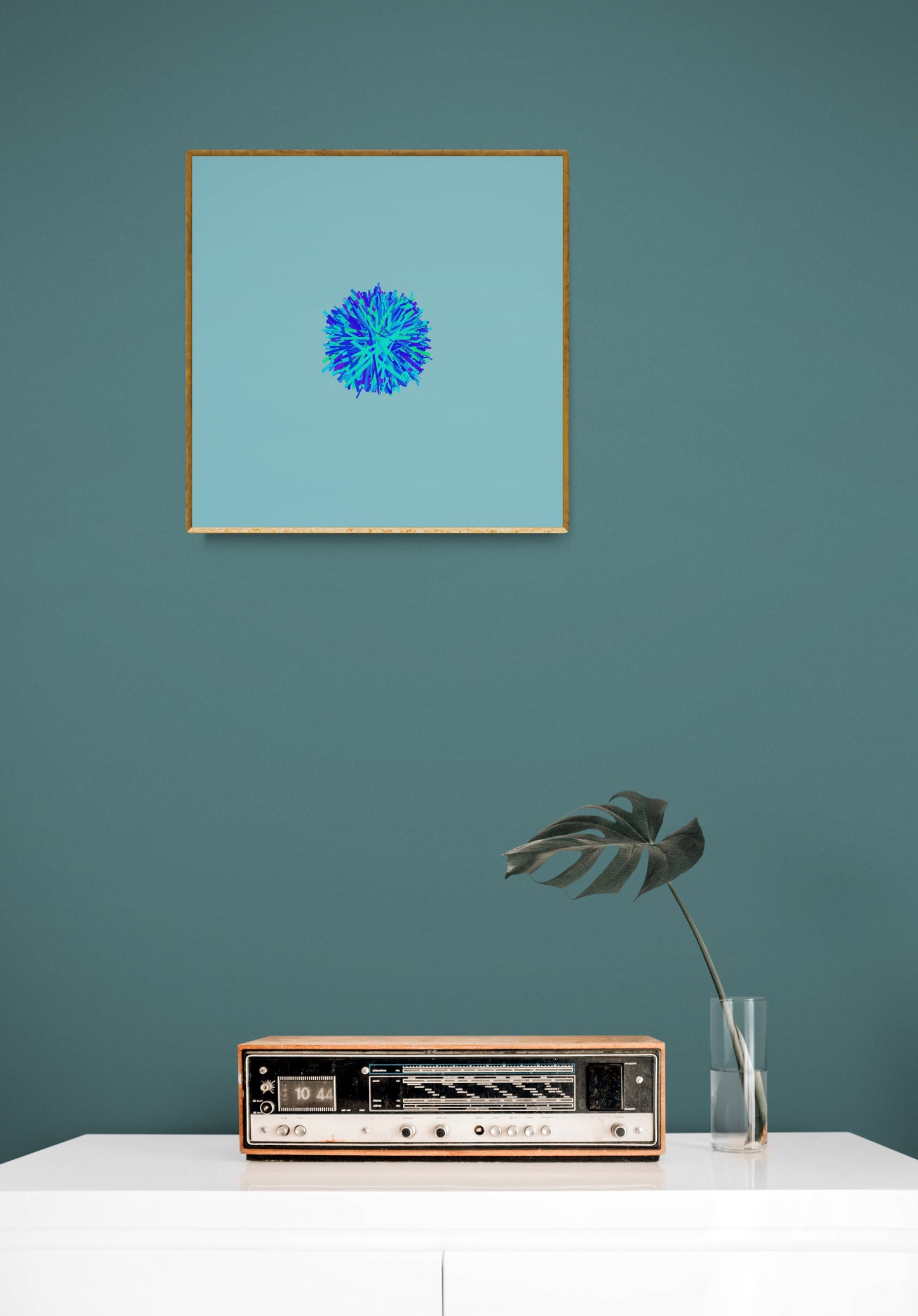 Framed abstract art print of blue, green and purple lines in a circular pattern on a light blue backgroundon a teal wall with a vintage radio and plant below.