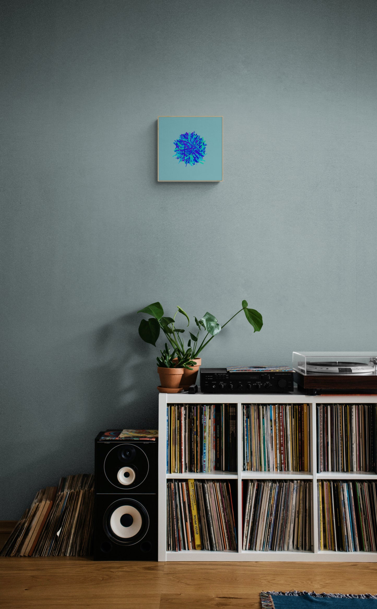 Room interior with vinyl records, speakers, and a plant against a gray wall and abstract art print of blue, green and purple lines in a circular pattern on a light blue background