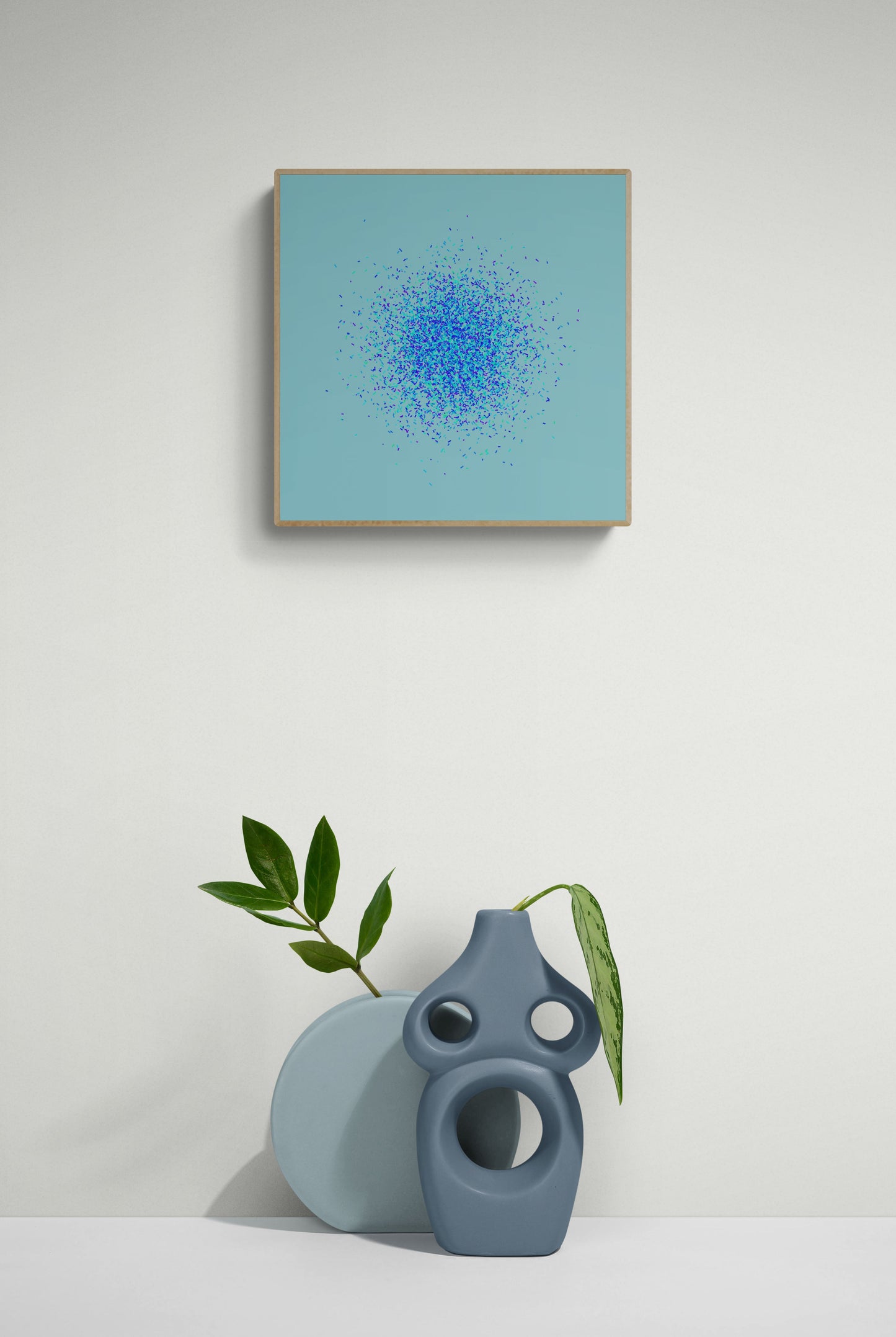 Framed abstract art print of blue, green and purple confetti like circular pattern on a light blue background on a white wall, accompanied by two ceramic vases.