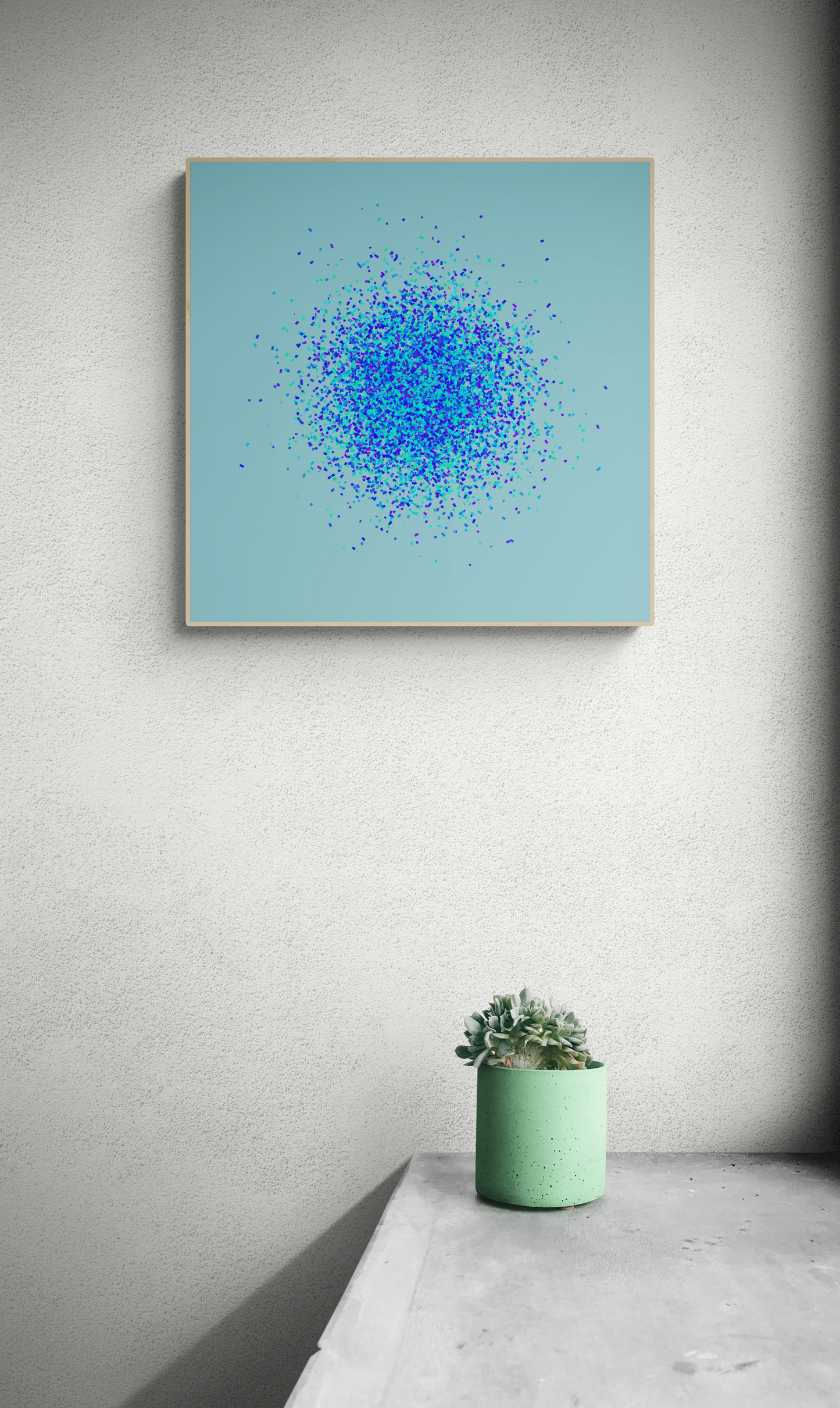 Framed abstract art print of blue, green and purple confetti like circular pattern on a light blue background on a light gray wall, below a small potted plant.