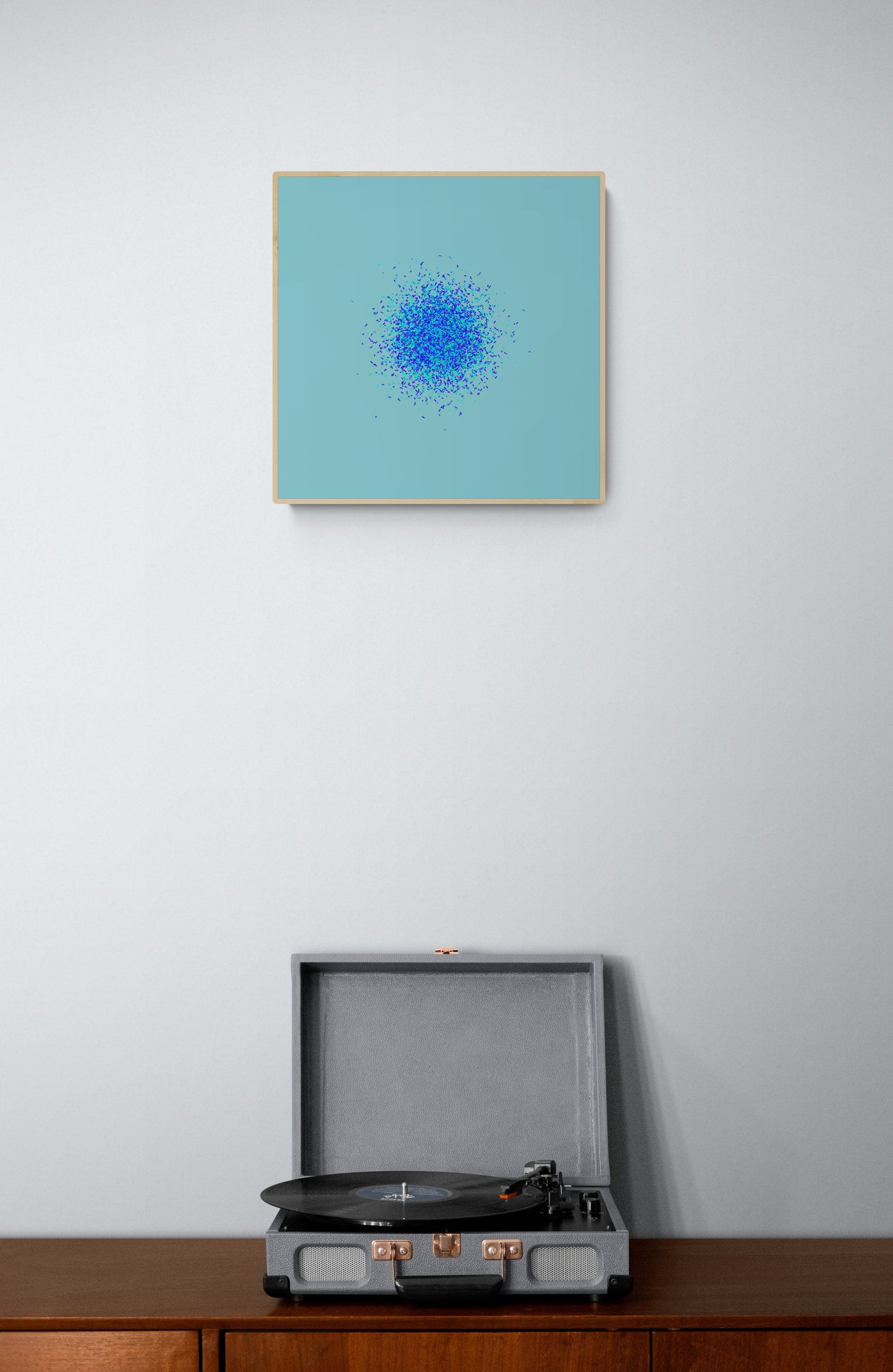 Framed abstract art print of blue, green and purple confetti like circular pattern on a light blue background on a wall above a record player on a wooden surface.