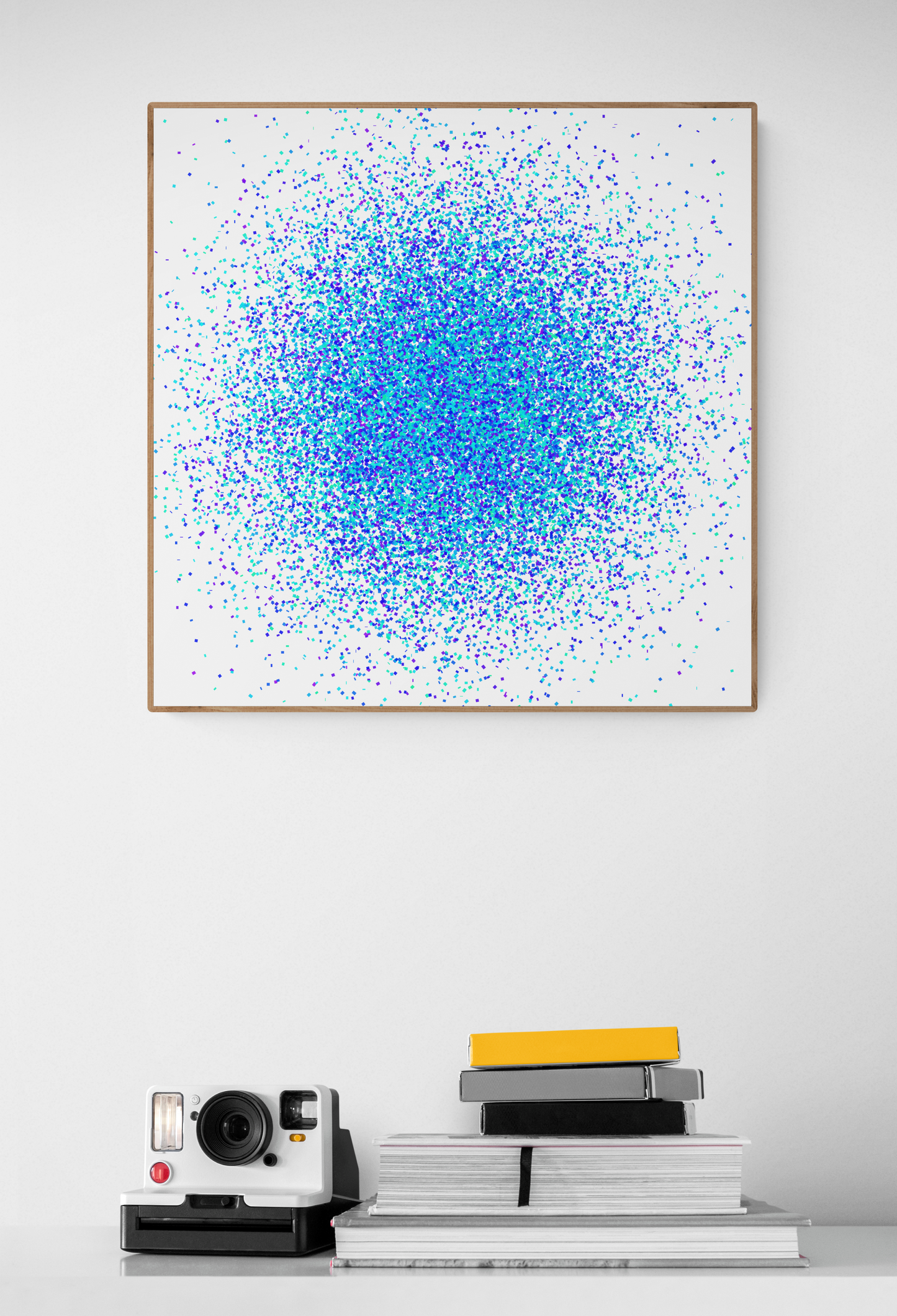 Framed abstract art print with blue, green and white dots on a white wall, below a stack of books and a camera.