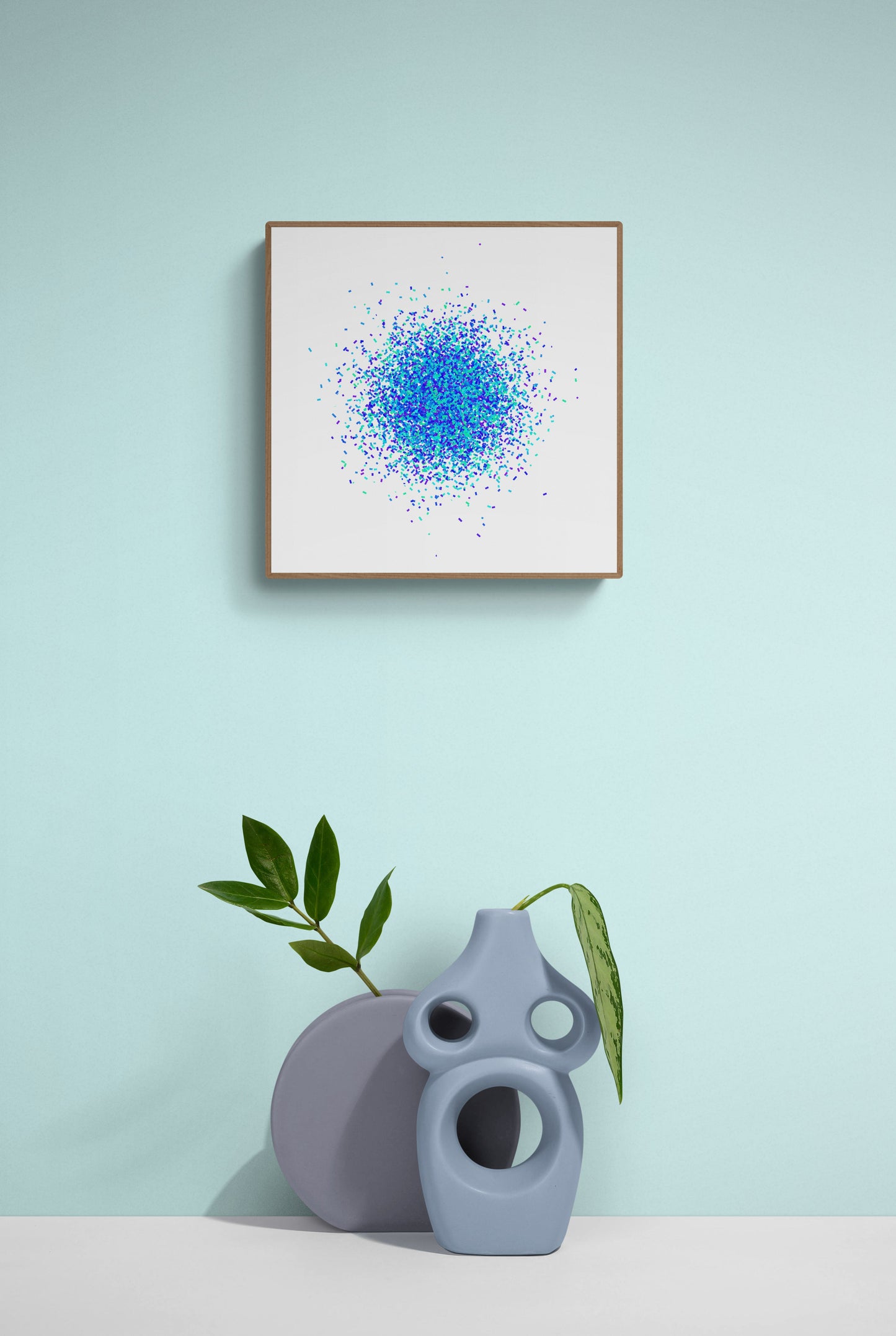 Framed abstract art print with blue and green pattern on a light blue wall, accompanied by a gray vase and plant.
