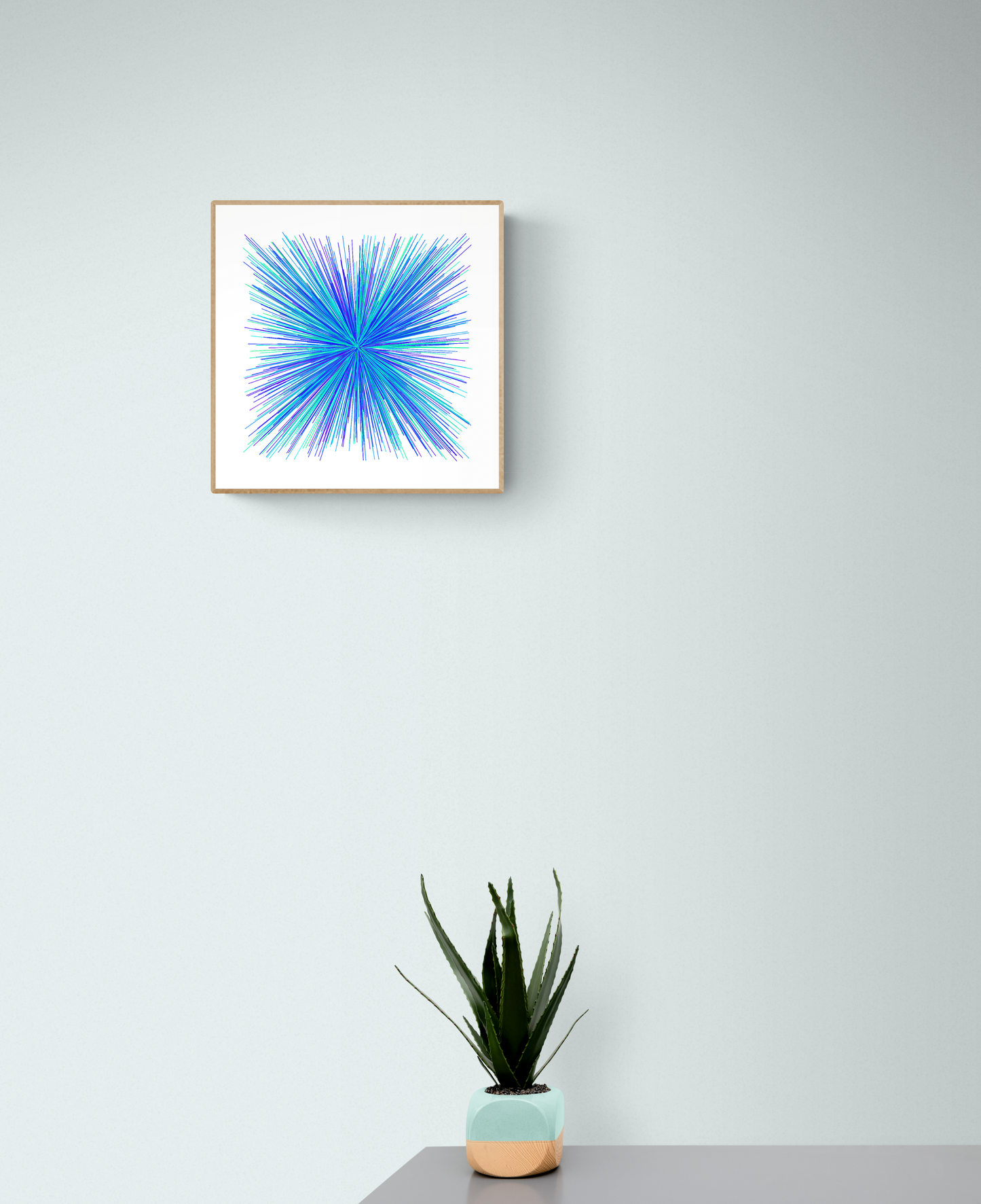 Framed abstract art print with blue, green and purple starburst pattern on a light gray wall, below a small potted plant.