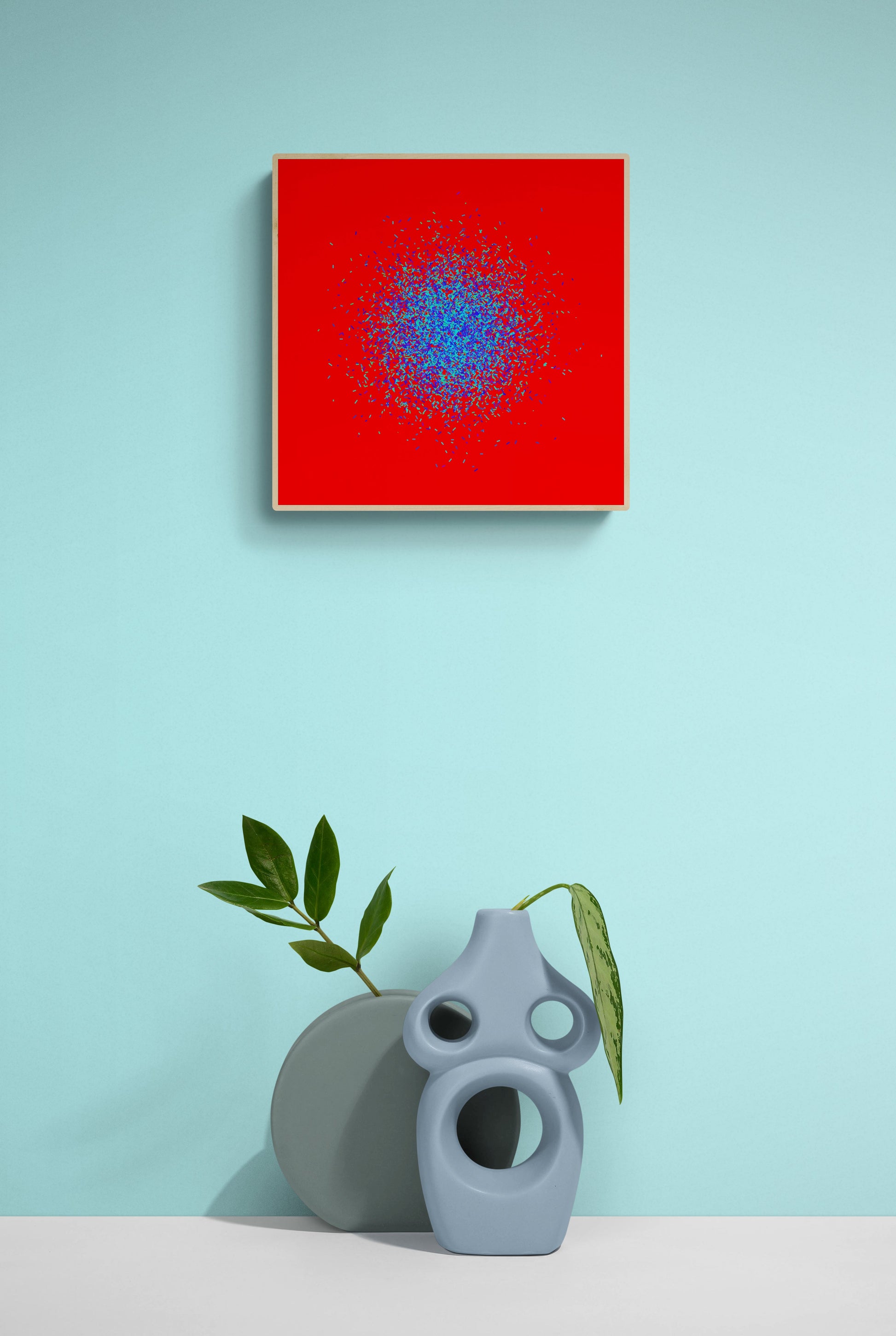 Blue, green, purple confetti like patterned abstract art print on a red background, placed above a gray vase and plant against a light blue wall.