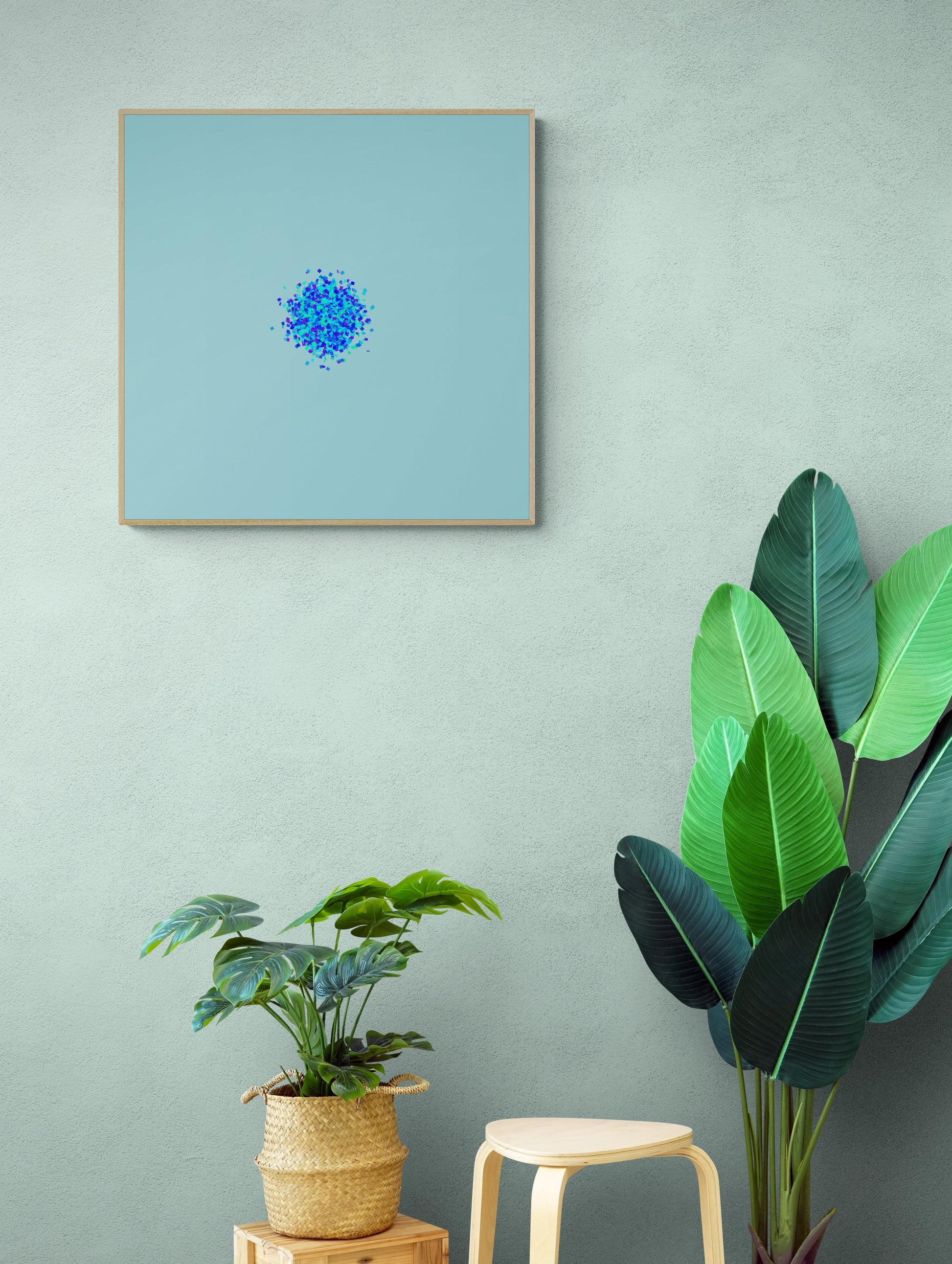 Framed abstract art print of blue, green and purple confetti like circular pattern on a light blue background on a wall with plants below